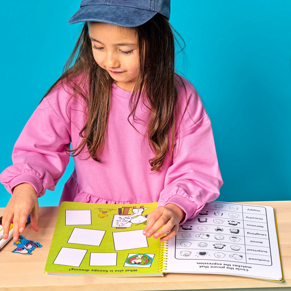 Child wearing a blue cap with a yellow patch, sitting at a table with educational materials against a blue background.
