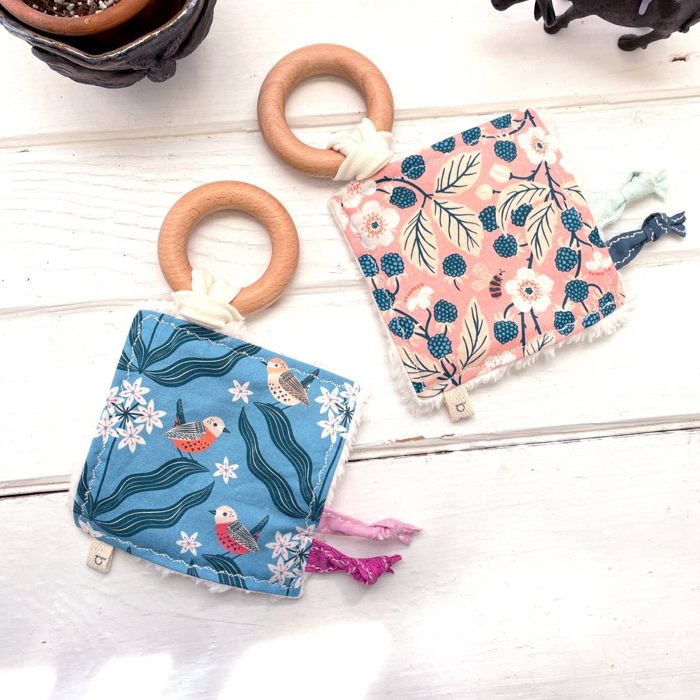 Two organic baby crinkle toys with a blue birds pattern and a pink berries and bees design, accompanied by a wooden teething ring, displayed on a white wooden surface.