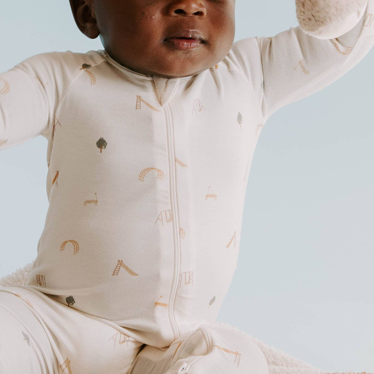 Baby in a beige onesie with play ground prints sitting on a textured blanket against a light blue background