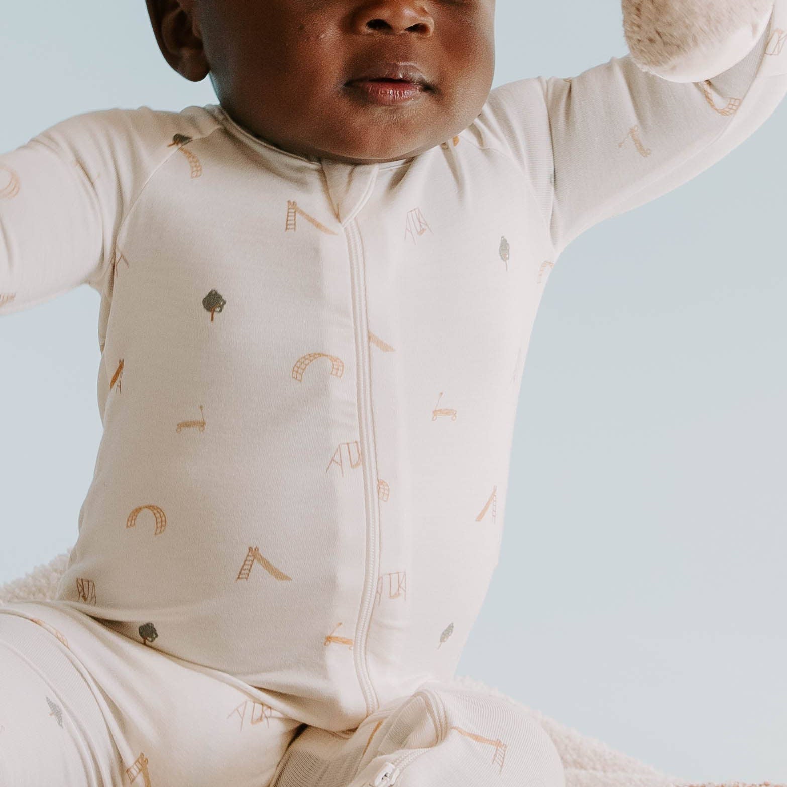 Baby in a beige onesie with play ground prints sitting on a textured blanket against a light blue background