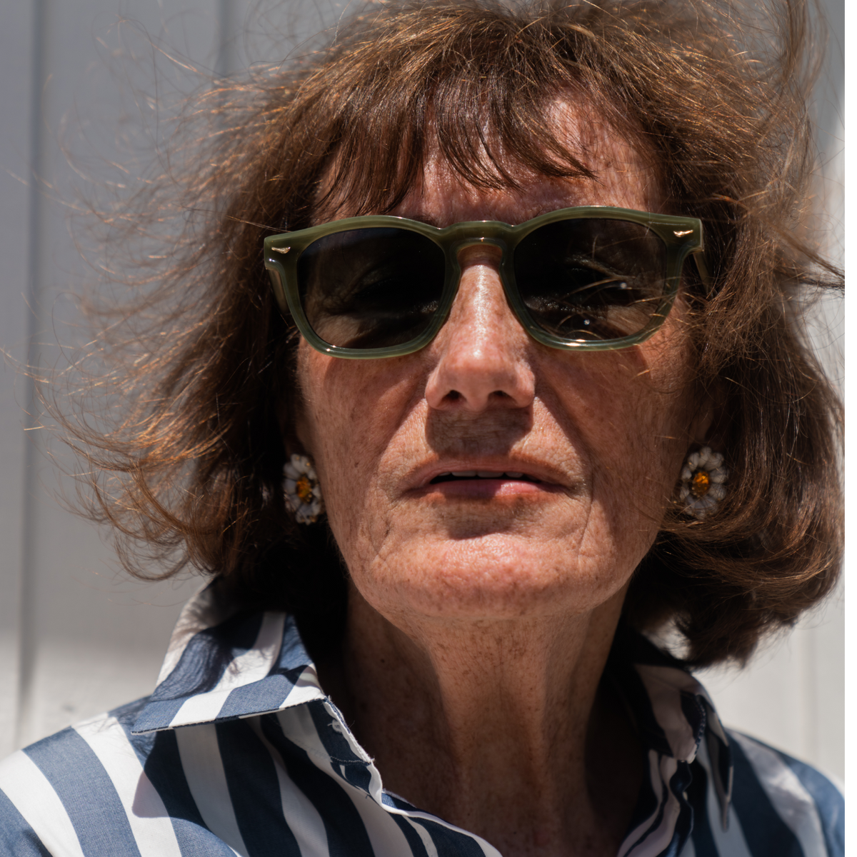 A close-up portrait of a woman with brown hair wearing dark green sunglasses and a blue and white striped button-up shirt, set against a light gray background.
