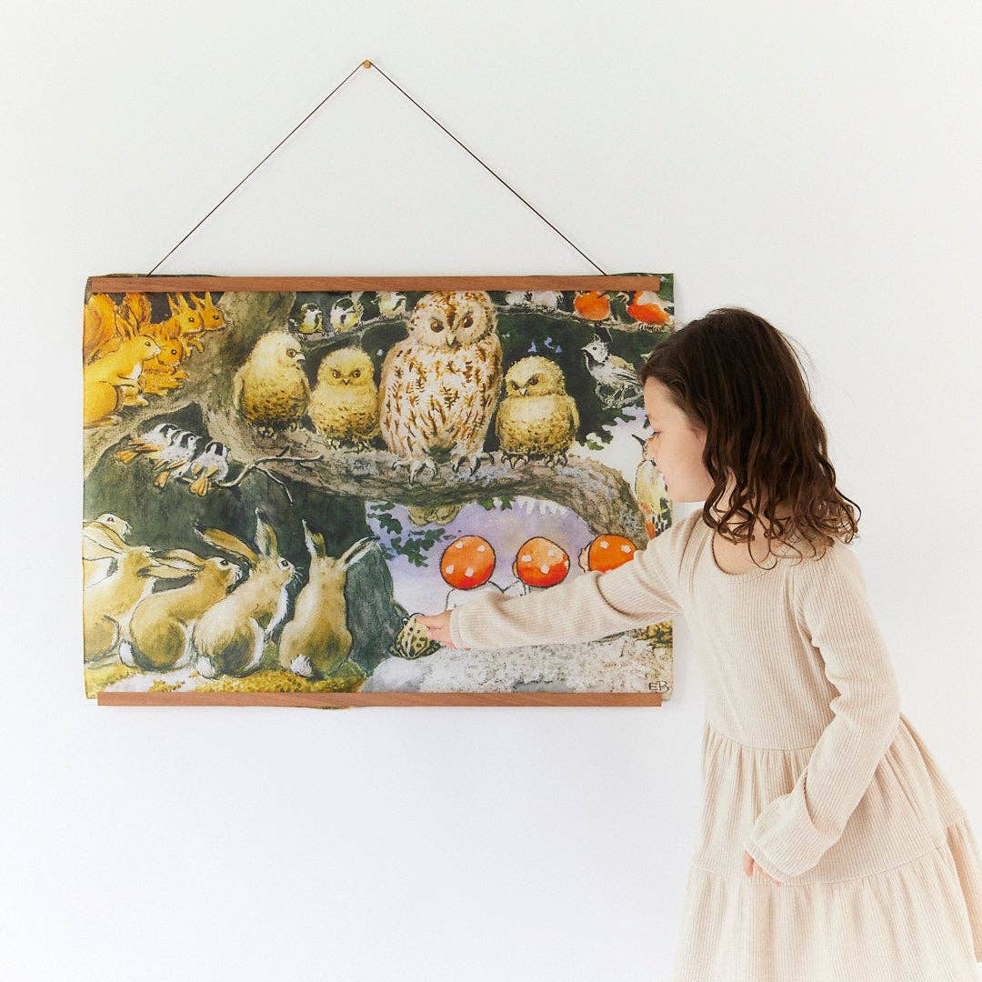 A child in a cream dress stands beside a large framed wall hanging depicting owls, rabbits, and red apples, photographed against a plain white wall.