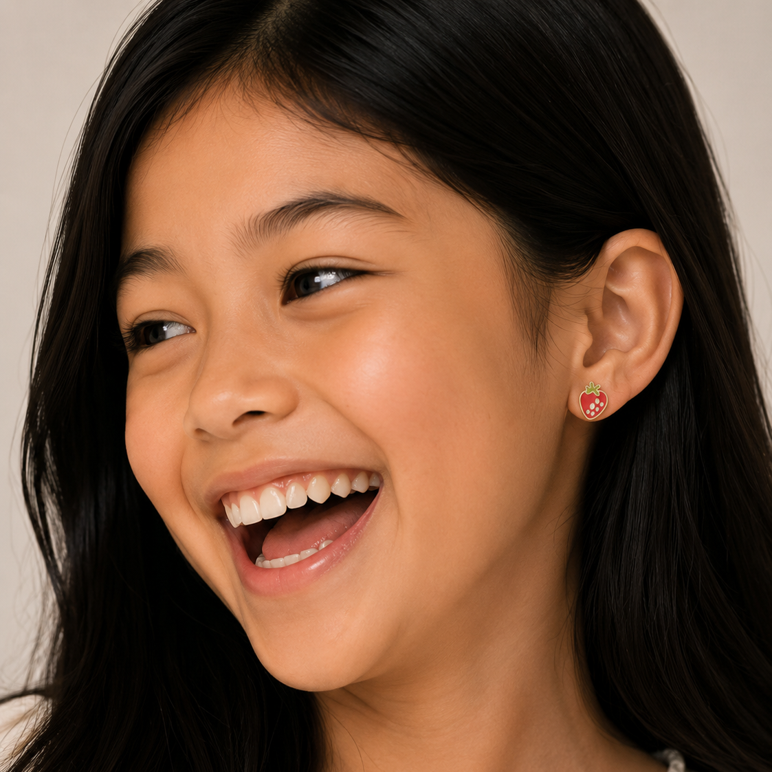 Young girl with a big smile wearing a strawberry-shaped earring against a neutral background