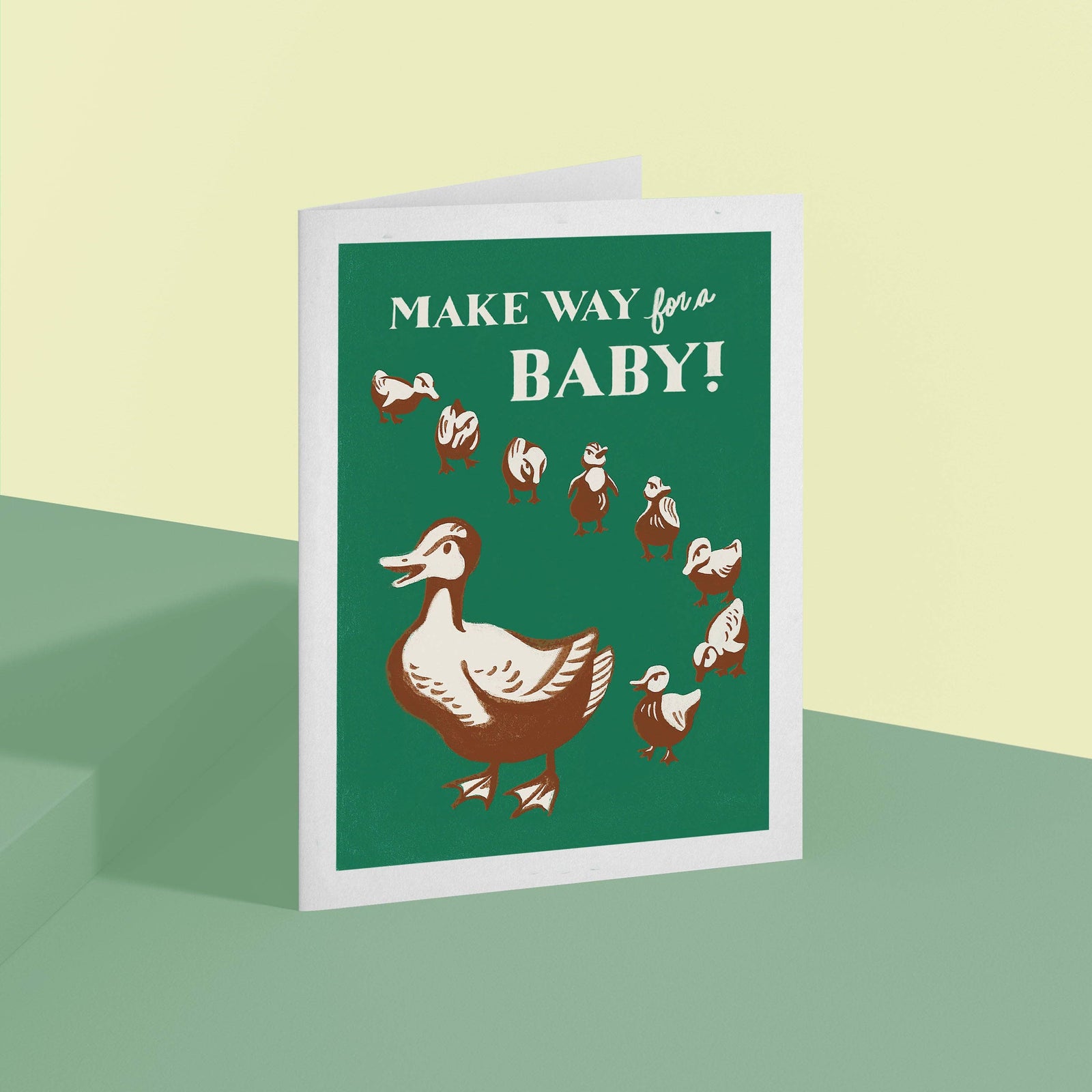 A greeting card with a green front featuring a brown duck and ducklings and the text 'MAKE WAY for a BABY!' in white, standing on a pale green surface against a pale yellow background.