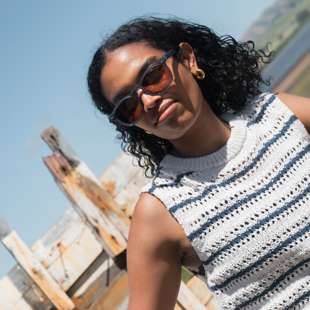 Woman wearing sunglasses and a sleeveless crochet top with a wooden structure and blue sky in the background