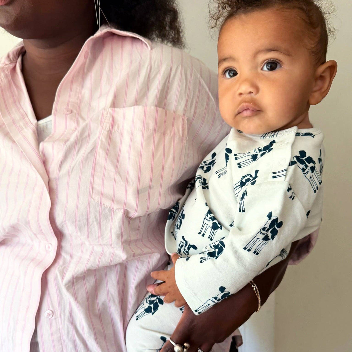 A baby wearing a white long-sleeve romper with a navy blue silhouette pattern of cows, held by an adult wearing a pink striped button-up shirt.