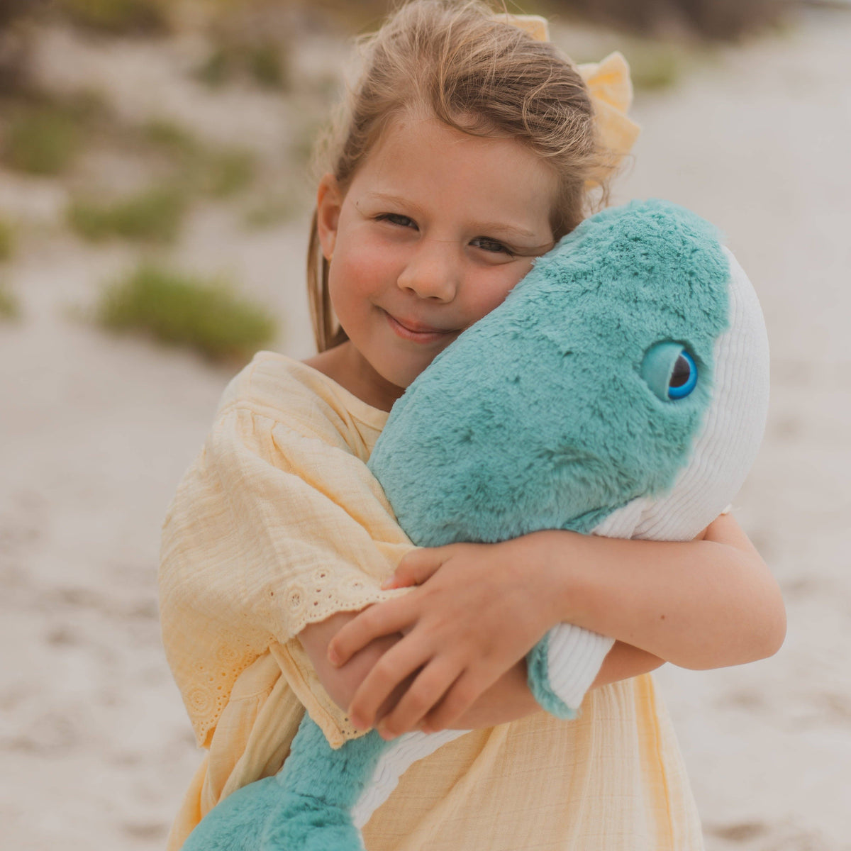 A young girl with blonde hair in a yellow dress hugs a large teal and white plush whale toy on a sandy beach.