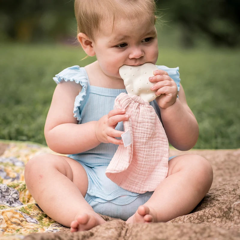 Baby in a light blue outfit with a pink teething toy sitting on a blanket outdoors.