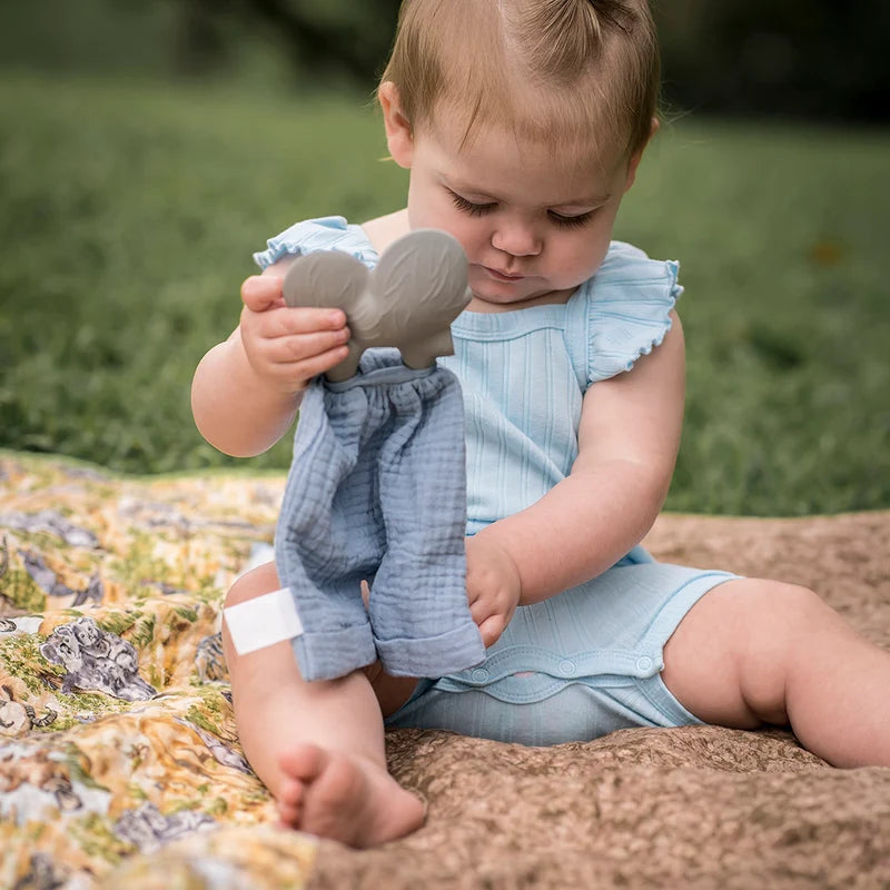 A baby sitting outdoors on a blanket wearing a light blue striped romper and holding a blue teething toy with blue fabric.