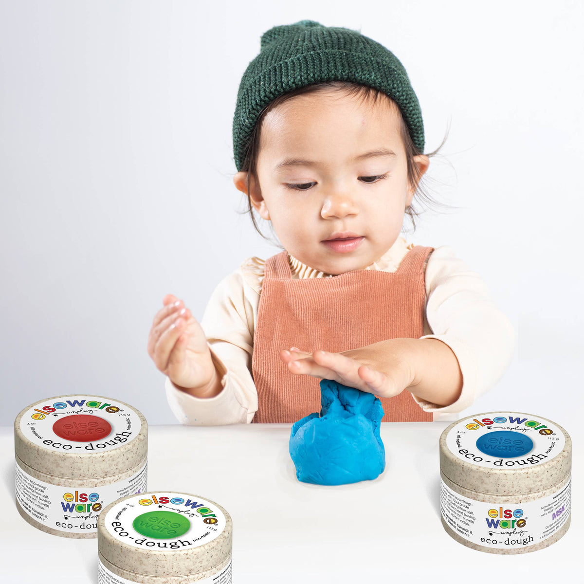 Child playing with blue play dough surrounded by three containers of play dough on a white background