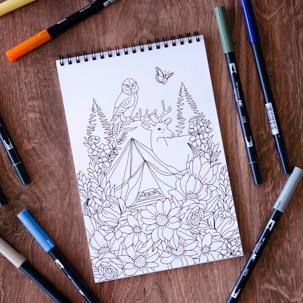Coloring book page with nature-themed design on a wooden surface with markers around.