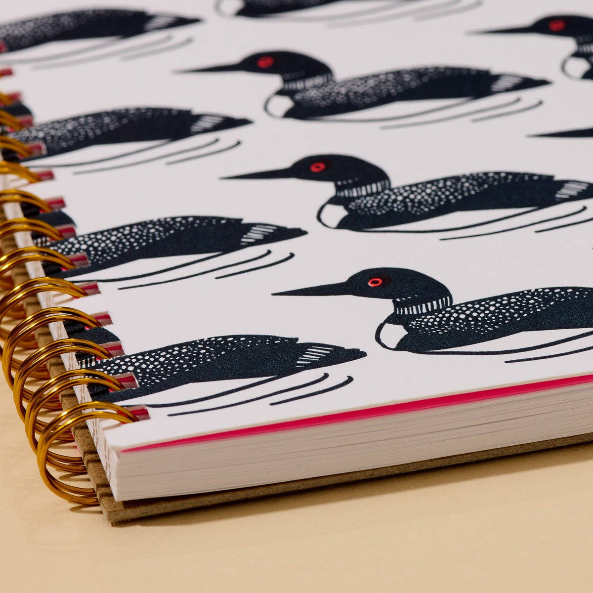 A close-up of a spiral-bound notebook with a white cover printed with black loon illustrations and small red eye accents, bound with a gold spiral on a beige surface.