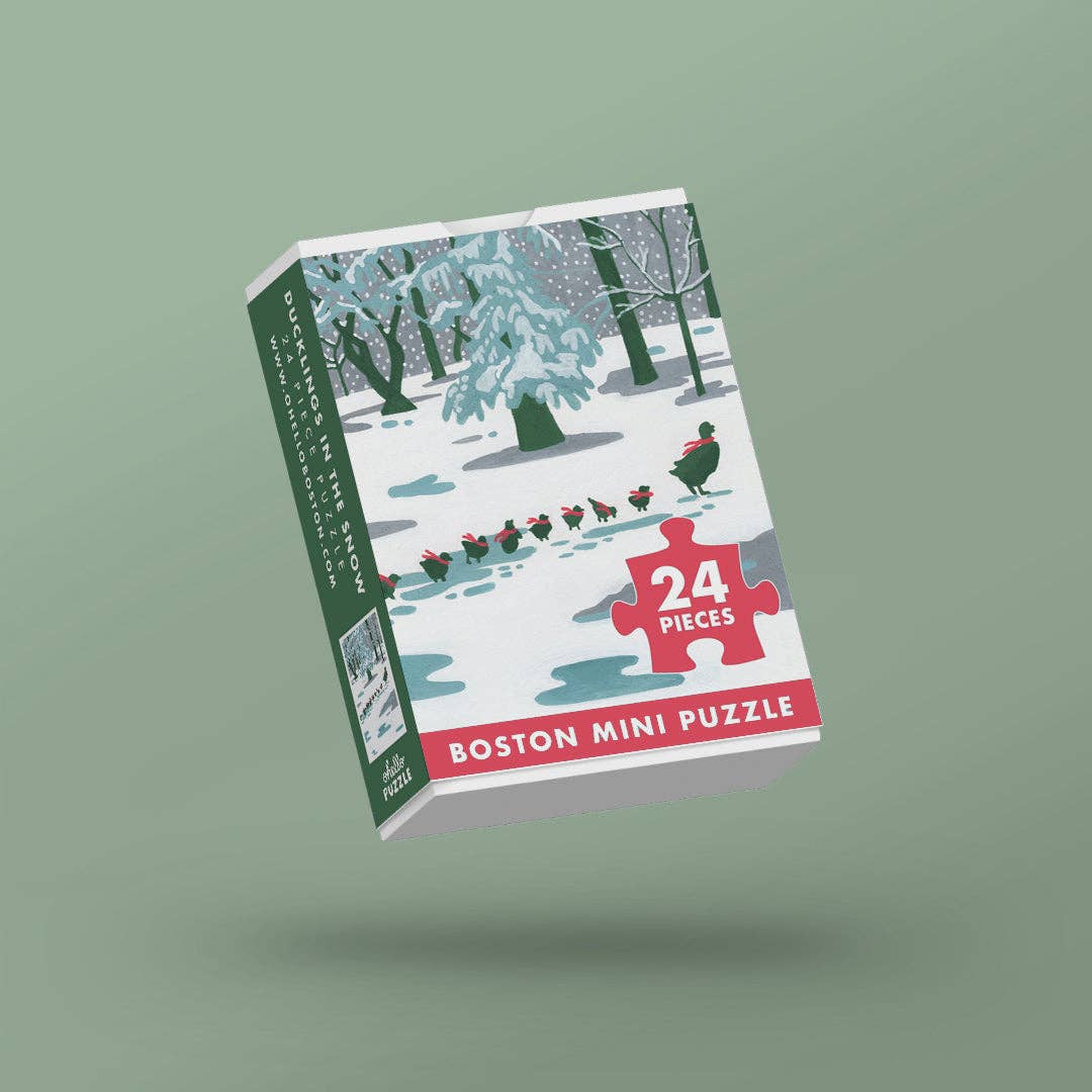Boston Mini Puzzle 24-piece jigsaw puzzle box featuring a winter forest illustration with snow-covered trees and a red puzzle-piece graphic indicating 24 pieces, shown floating against a muted sage green background.