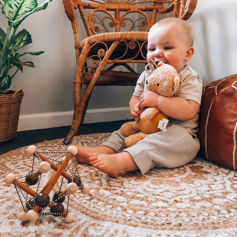 Baby holding a teddy bear in a room with a wicker chair and decorative items.