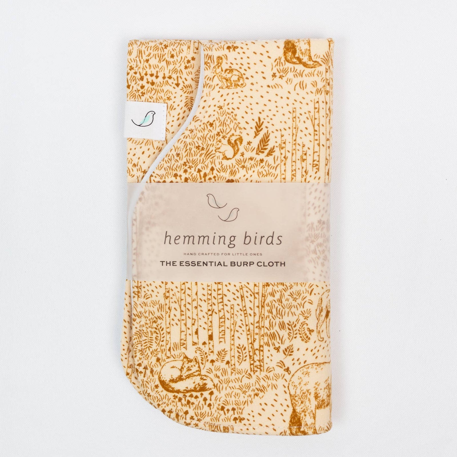 A beige burp cloth with a woodland animal pattern, rolled up and secured with a tie, labeled with 'hemming birds' and 'THE ESSENTIAL BURP CLOTH'.