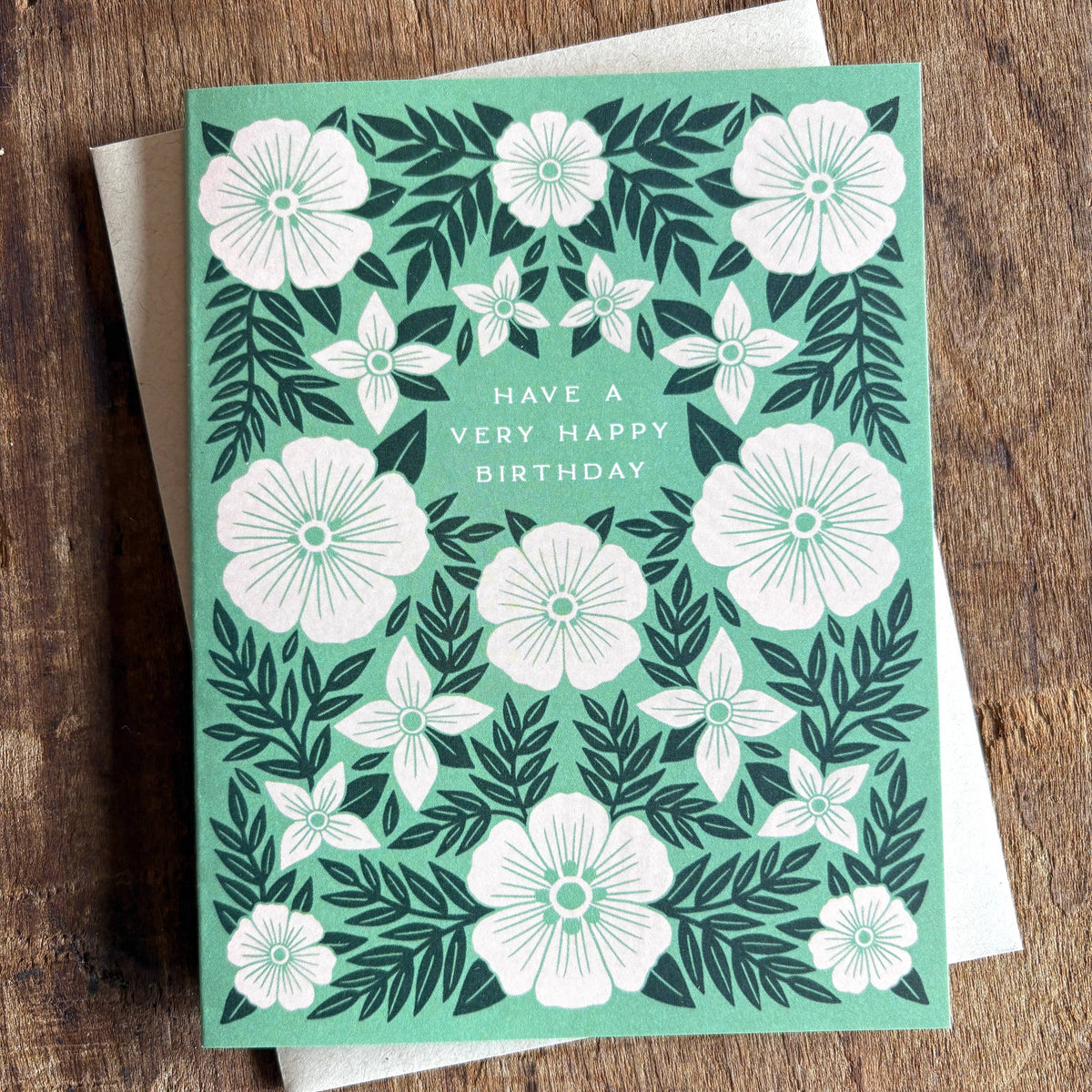 A square birthday card with a mint green background and white floral pattern, reading 'HAVE A VERY HAPPY BIRTHDAY' in the center, placed on top of a white envelope over a rustic wooden surface.