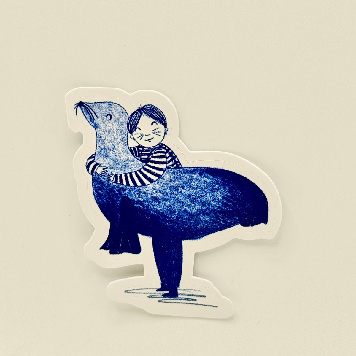 A die-cut sticker featuring a whimsical illustration of a child with glasses riding on the back of a large blue seal, shown against a plain beige background.