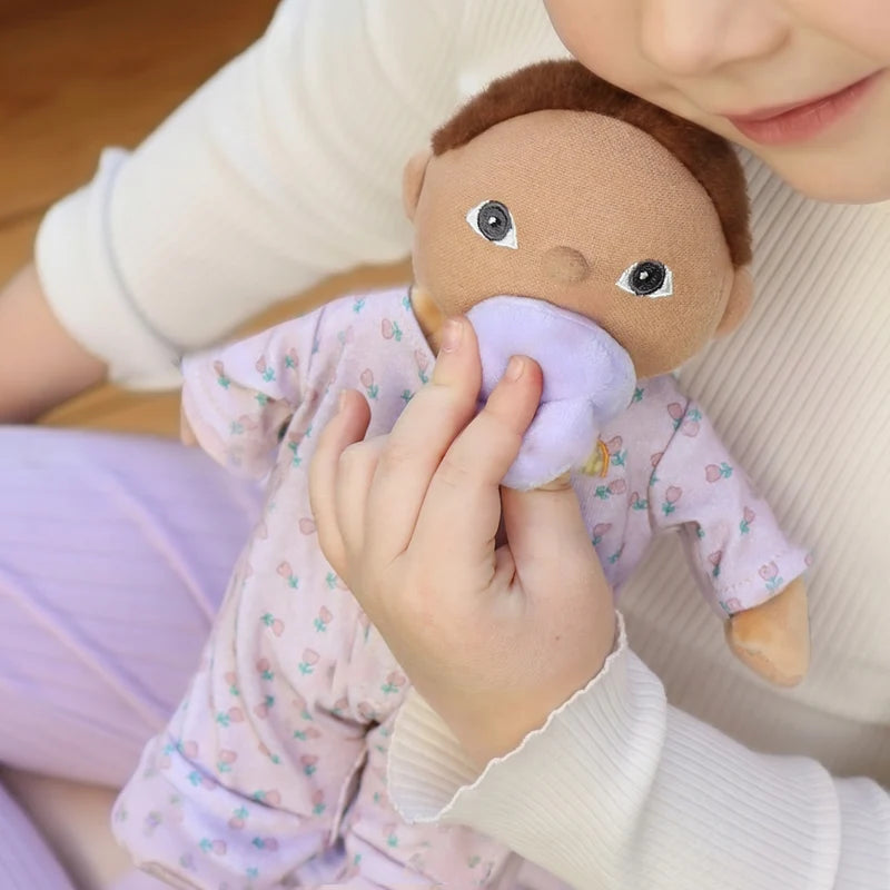 A baby doll with brown hair and a pacifier, dressed in a light pink patterned onesie, displayed inside a matching light pink bassinet bag. A small matching pacifier bottle is placed to the lower left, all on a clean white background.