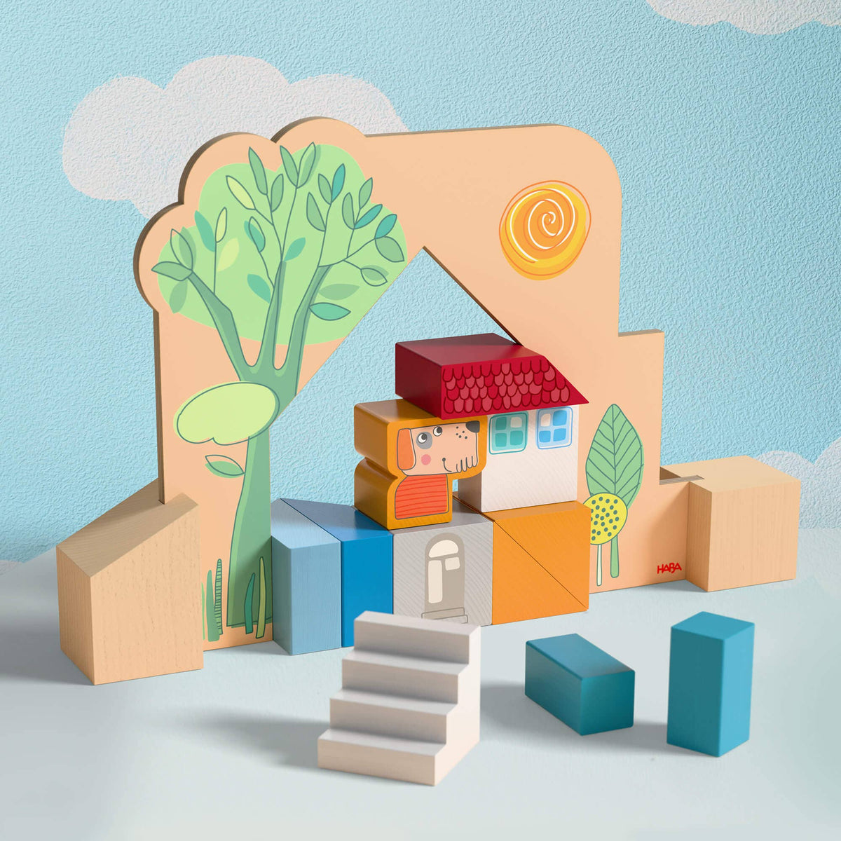 A children's wooden toy set featuring a house-shaped backdrop with a tree illustration, colorful stacked blocks, small steps, and geometric pieces on a light blue surface against a blue cloud-patterned background.