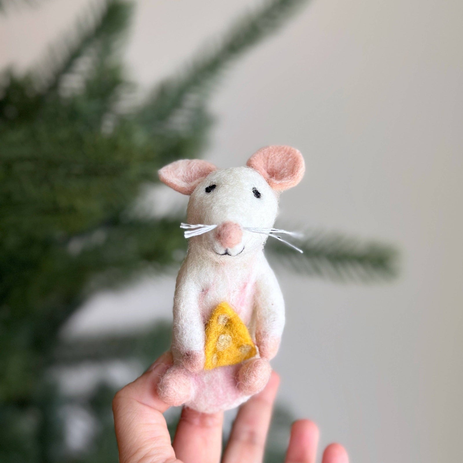 A hand holding a small felted wool mouse figurine with pink ears and feet, a white body, black eyes, white whiskers, and a yellow triangular cheese piece in its front paws, against a soft-focus background of green pine branches and a light wall.