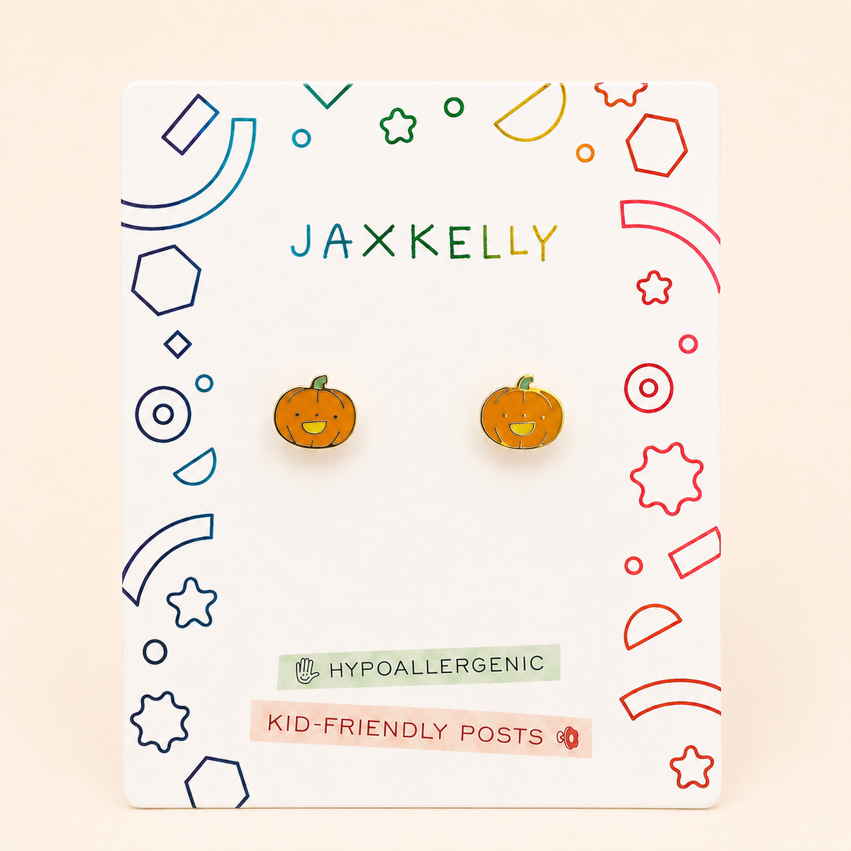 A pair of orange pumpkin-shaped stud earrings with green stems and yellow smiling faces, displayed on a white JAXKELLY brand card with colorful geometric borders.