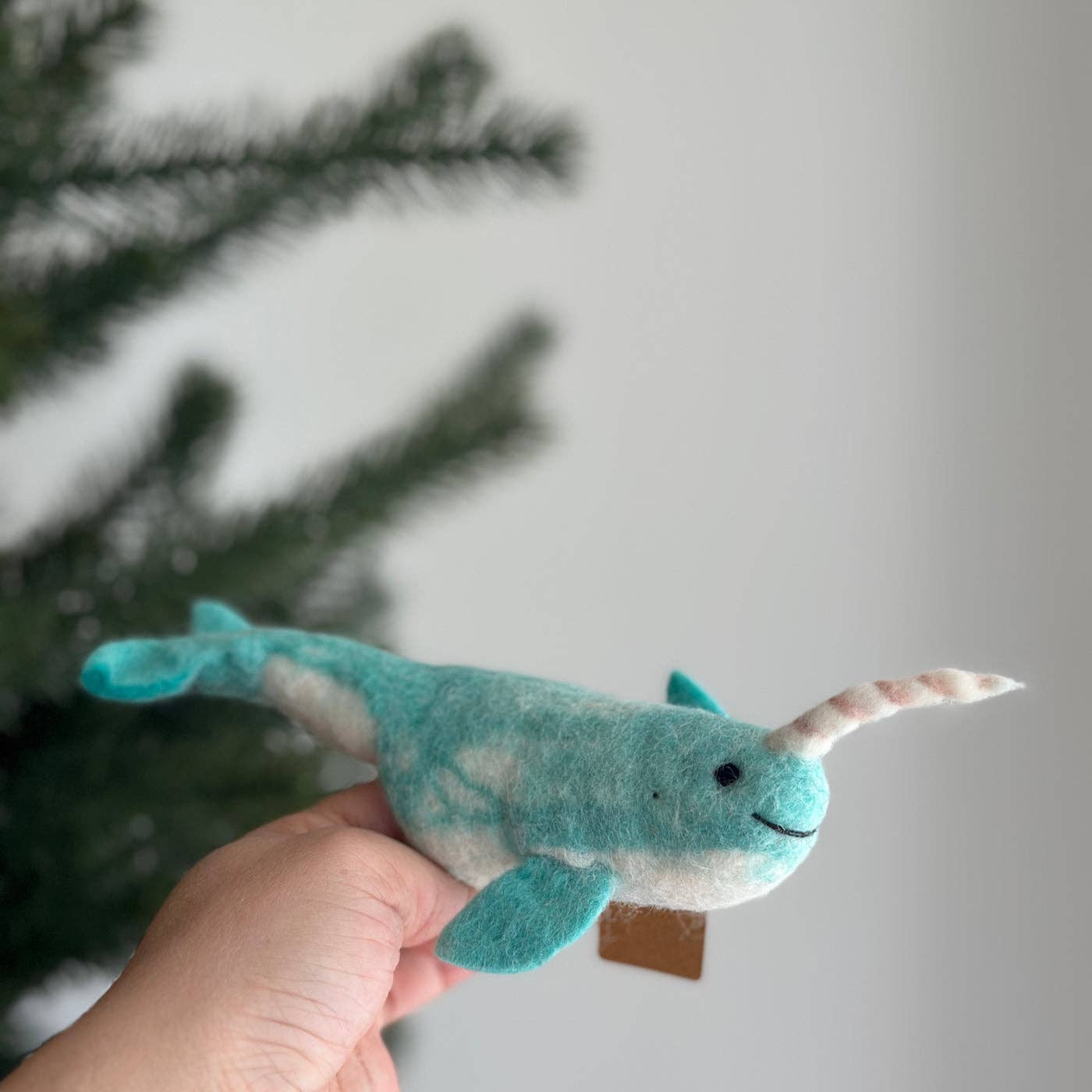 Plush toy narwhal held in a hand with a blurred Christmas tree background