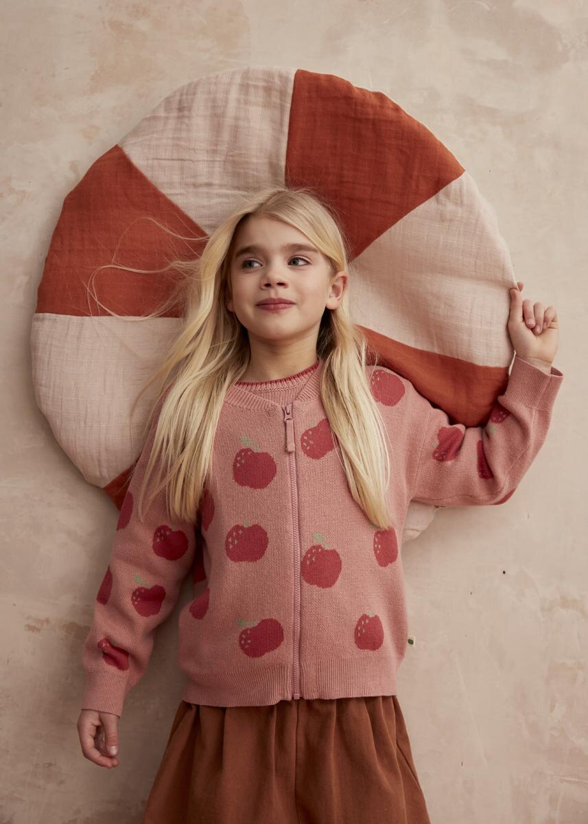 Young girl wearing a pink sweater with red  apple patterns, standing against a textured wall with a large circular pillow.