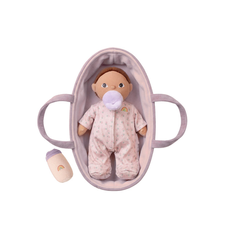A baby doll with brown hair and a pacifier, dressed in a light pink patterned onesie, displayed inside a matching light pink bassinet bag. A small matching pacifier bottle is placed to the lower left, all on a clean white background.