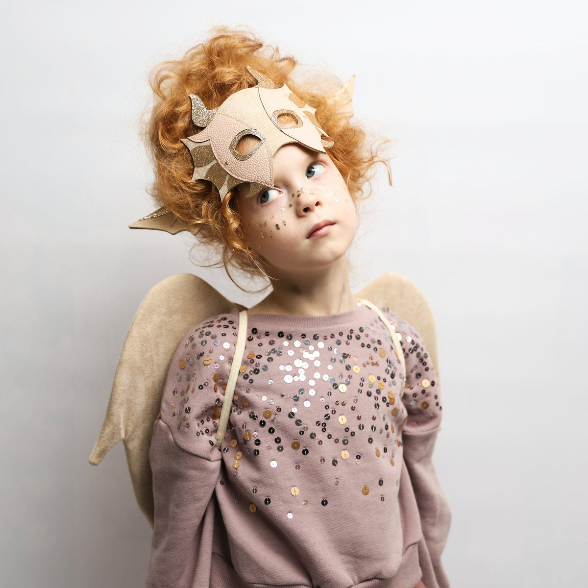 A child wearing a beige angel costume set, including a horned mask, pointed ears, and fabric wings, paired with a sparkly mauve sweatshirt, against a plain light gray background.