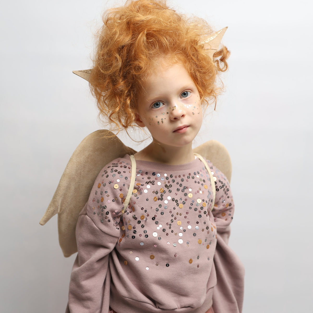 A child wearing a pink sequined long-sleeve top with dragon wings and pointed ears, posed against a plain light gray background.