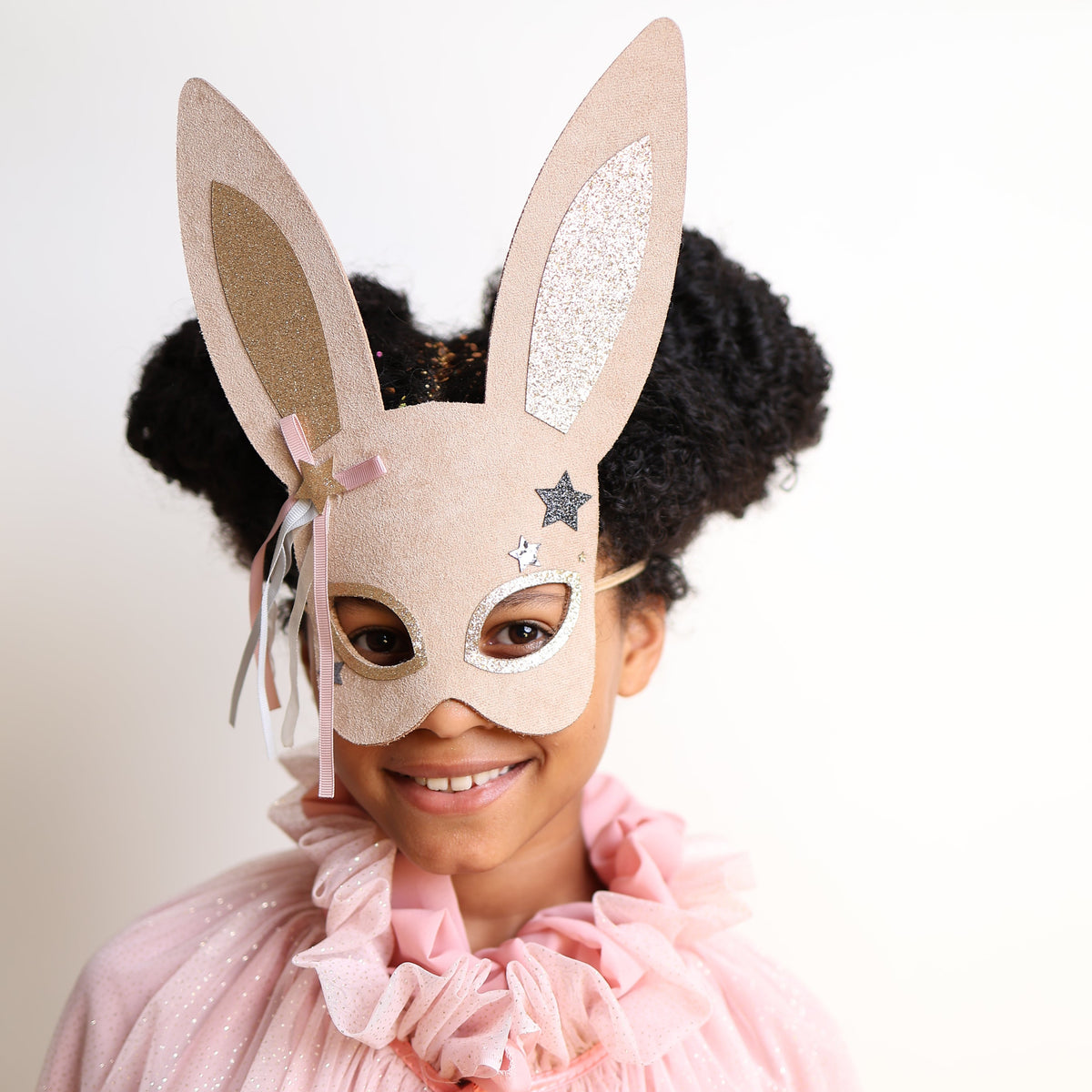 A young girl with dark curly hair wearing a beige bunny mask with glittery ears and a pink ruffled cape, smiling against a plain light background.