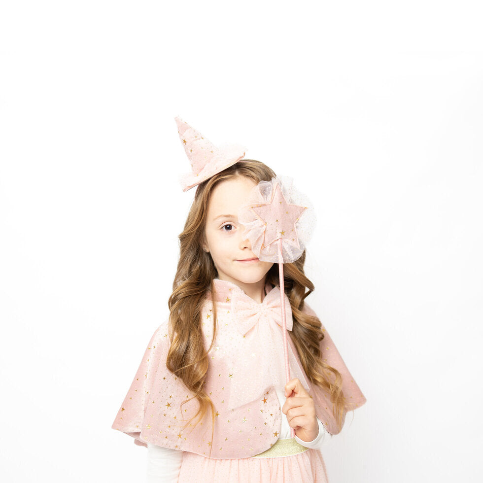 A young girl with long wavy brown hair wearing a pink sparkly costume cape and matching pointed hat, holding a star-shaped wand over her eye against a plain white background.