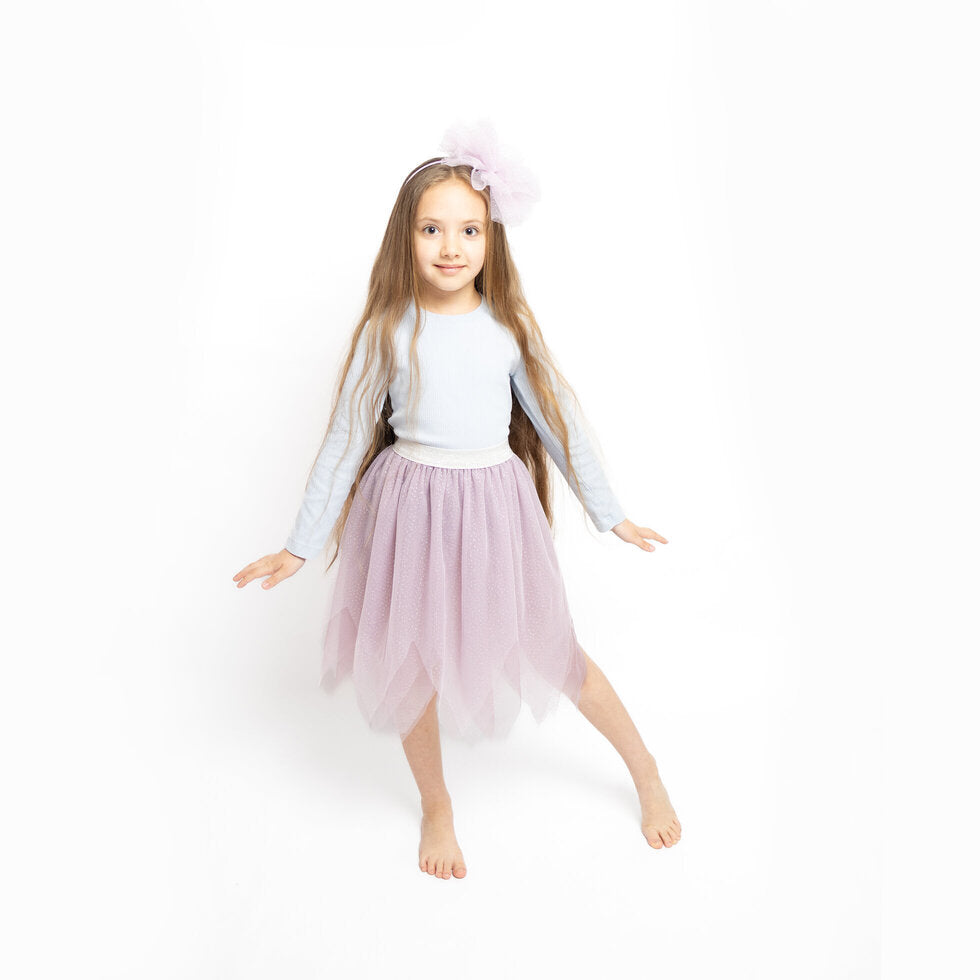 A young girl with long light brown hair stands barefoot against a white studio background, wearing a white long-sleeve top, a light pink tulle tutu skirt, and a matching pink bow headband.