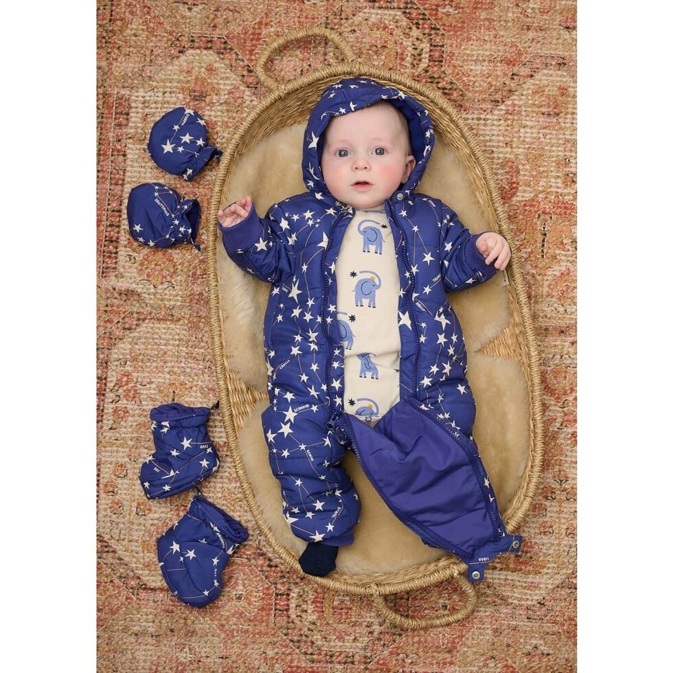 Baby in a blue star-patterned snowsuit lying on a white cushion against a beige background