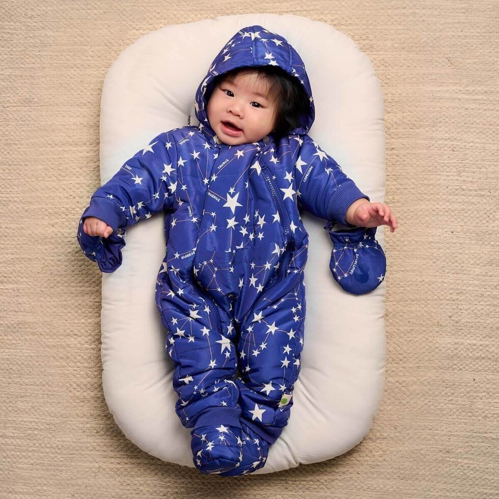 Baby in a blue star-patterned snowsuit lying on a white cushion against a beige background