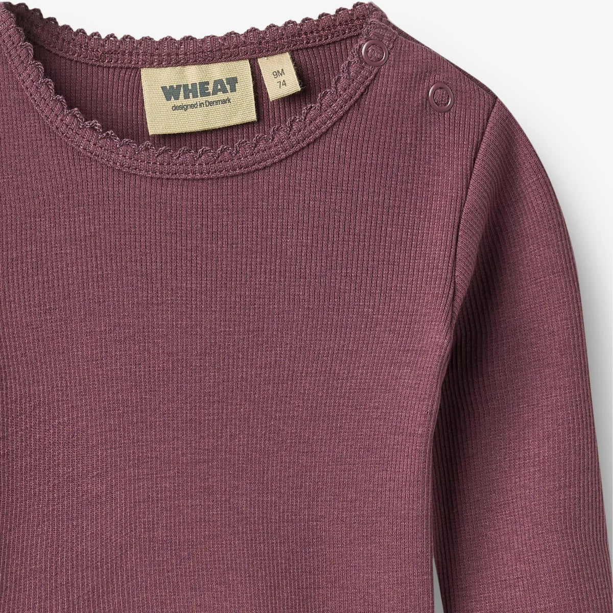 Close-up of a ribbed knit long-sleeve top in dusty mauve, showing the crew neckline with scalloped trim, shoulder buttons, and interior labels reading WHEAT 