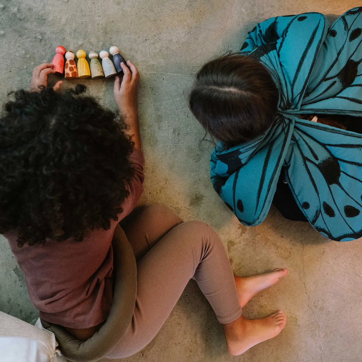 Two children sitting on the floor with a collection of colorful peg dolls and a blue butterfly-shaped costume
