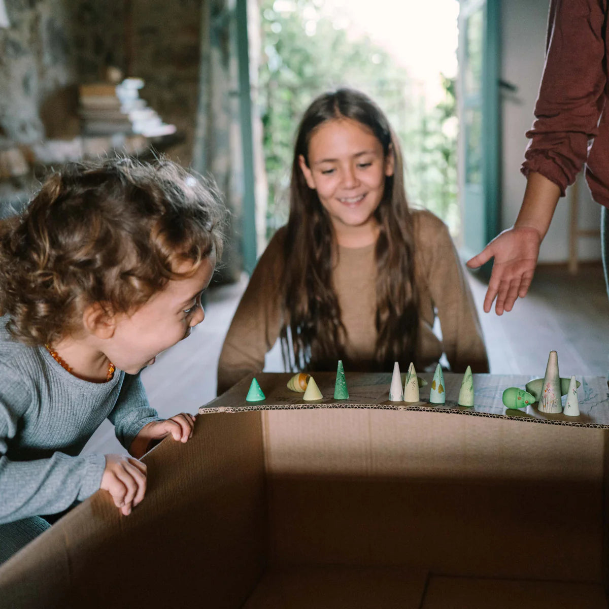 Two children playing with a cardboard box toy resembling a forest, with an adult's hand visible.