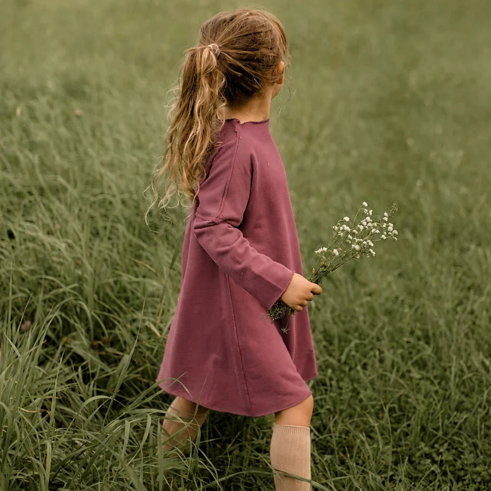 A young girl with a ponytail stands in a grassy field, viewed from behind, wearing a dusty rose long-sleeve dress and holding a small bunch of white flowers.