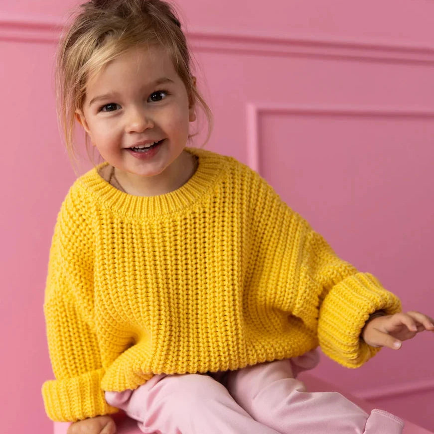 Child wearing a yellow sweater and pink pants sitting on a pink block against a pink background