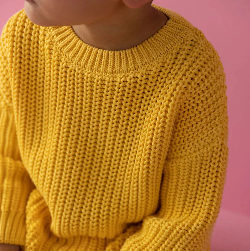 Child wearing a yellow sweater and pink pants sitting on a pink block against a pink background