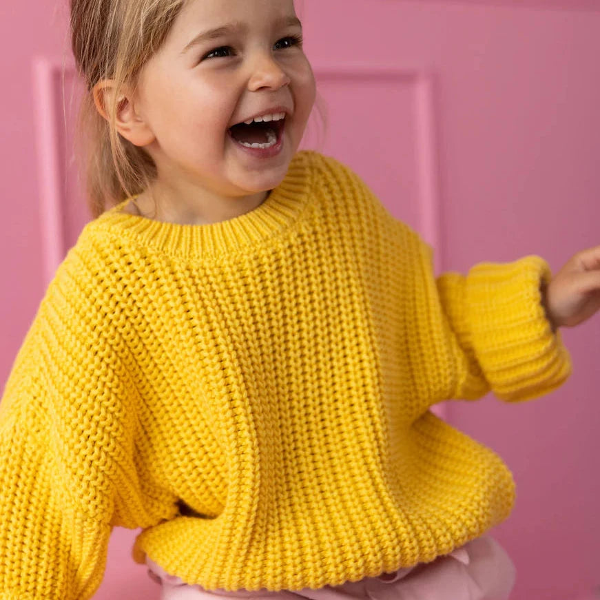 Child wearing a yellow knitted sweater and pink pants against a pink background