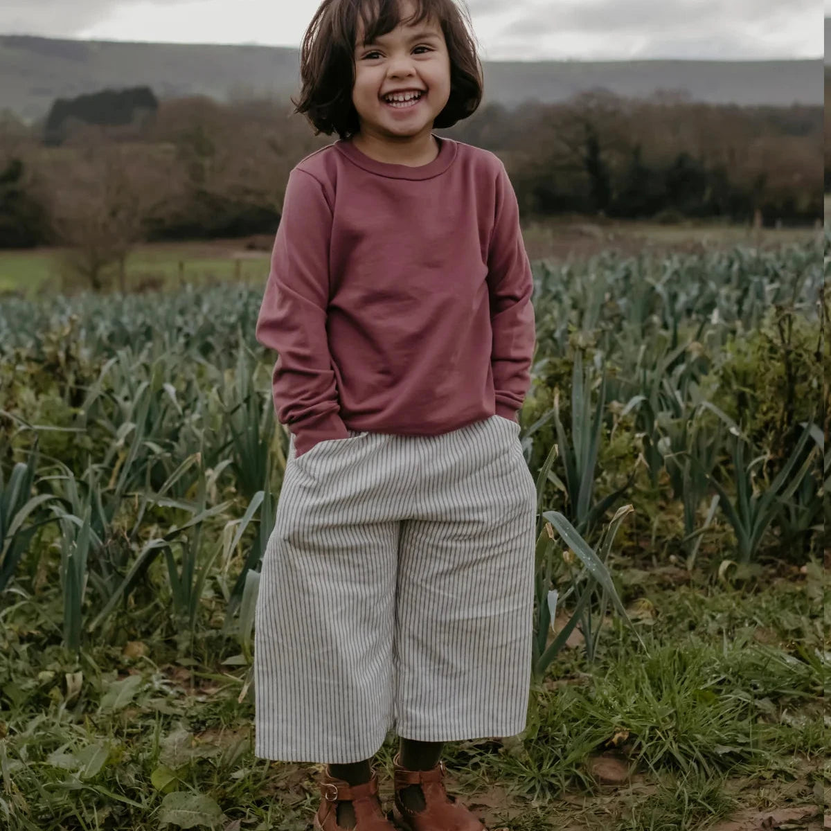 A young child standing outdoors in a field, wearing a dusty rose long-sleeve top, wide-leg light gray and white striped trousers, and brown boots.