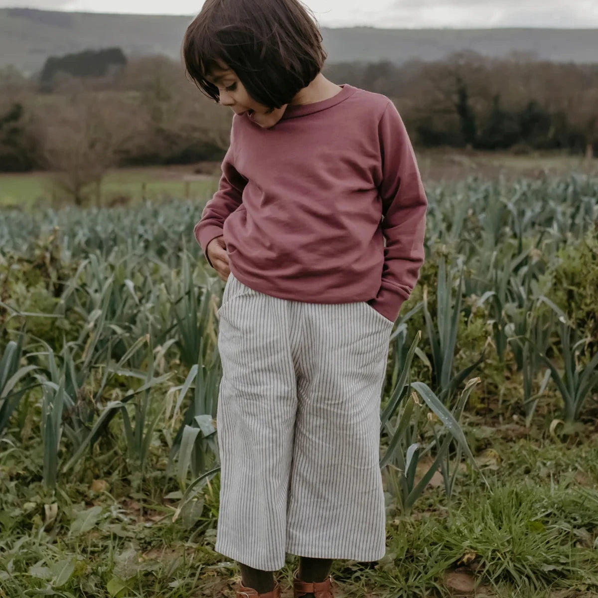 A child standing in a field wearing a dusty rose long-sleeve top, white and gray vertically striped cropped trousers, and brown leather boots.
