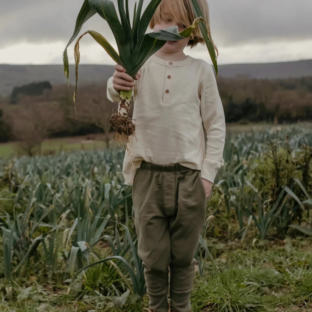 A young child standing in a field outdoors, holding a large green plant over their face. The child is wearing a cream long-sleeve henley shirt, olive green pants, and brown leather shoes under an overcast sky.