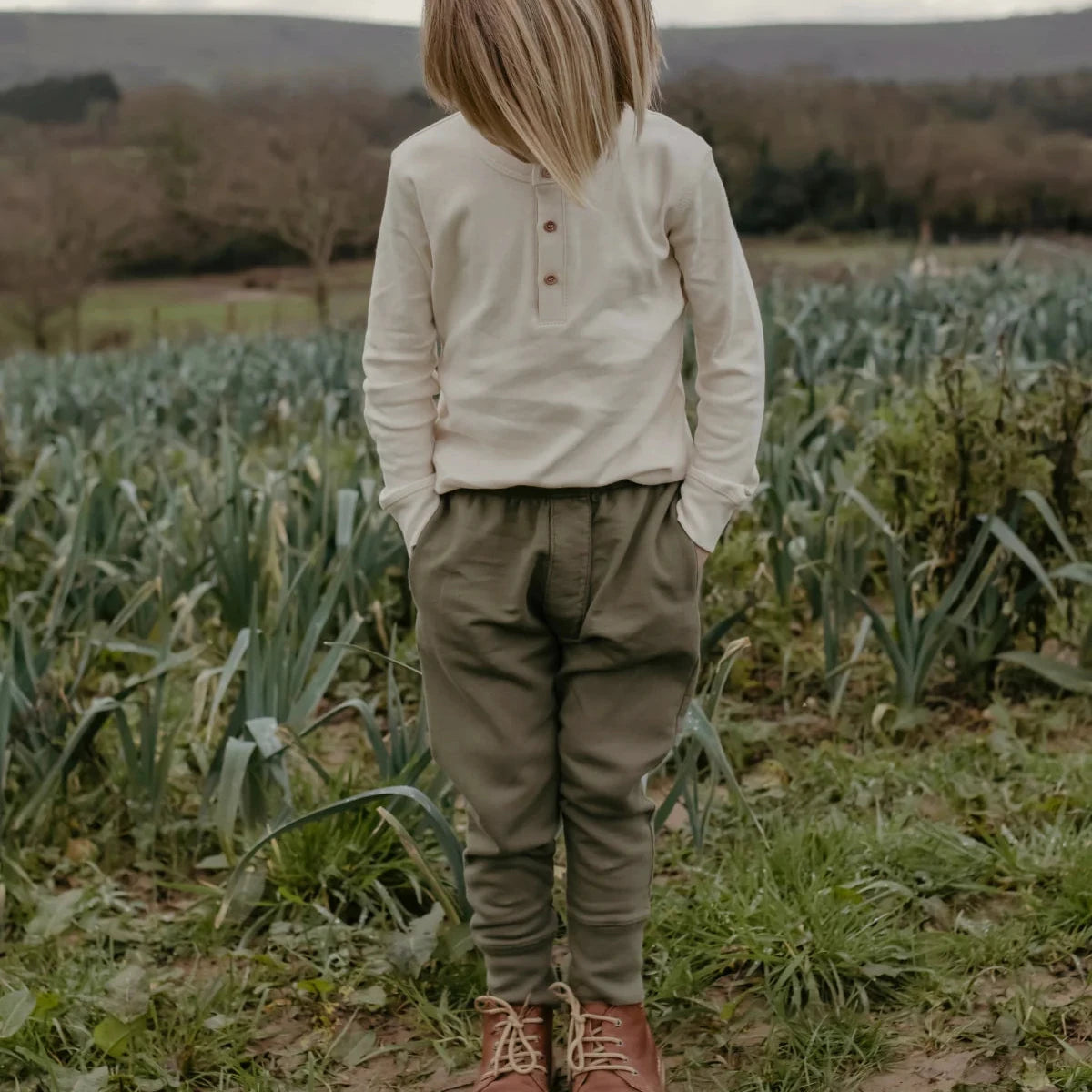 A child standing outdoors in a field, viewed from behind, wearing a cream long-sleeve henley shirt, olive green trousers, and brown lace-up boots under an overcast sky.