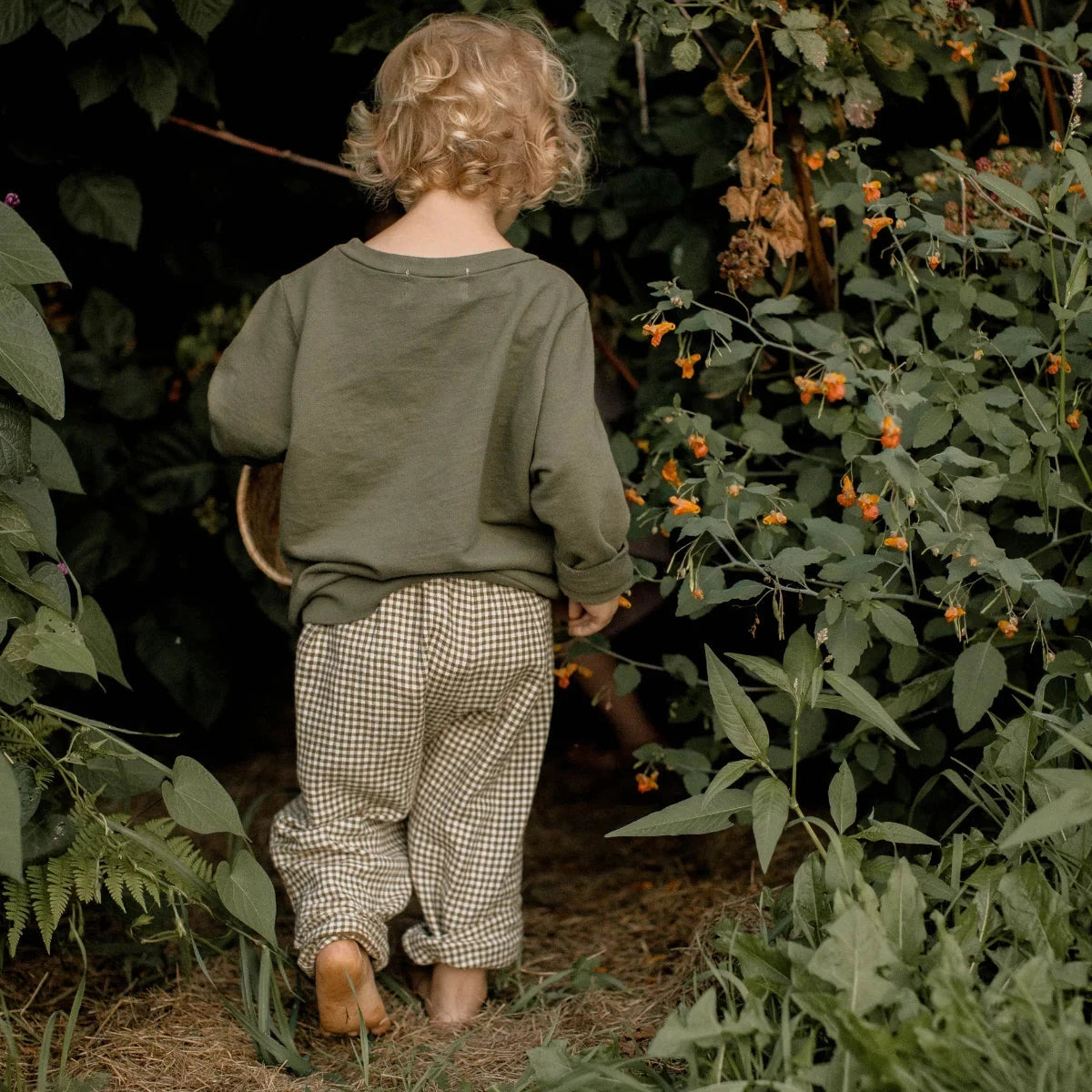 A child seen from behind, wearing an olive green long-sleeve top and beige-and-brown gingham pants, walking barefoot through a lush garden path surrounded by green foliage and small orange flowers.