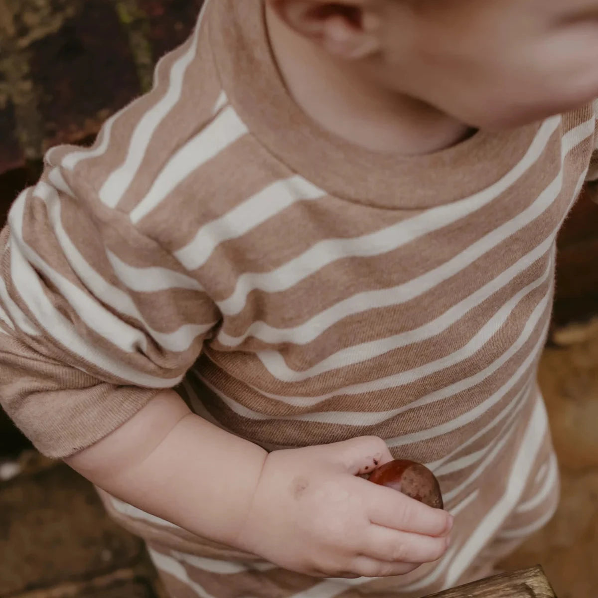 A close-up lifestyle shot of a toddler wearing a long-sleeve knit top with horizontal brown and cream stripes, holding a small acorn in a rustic setting.