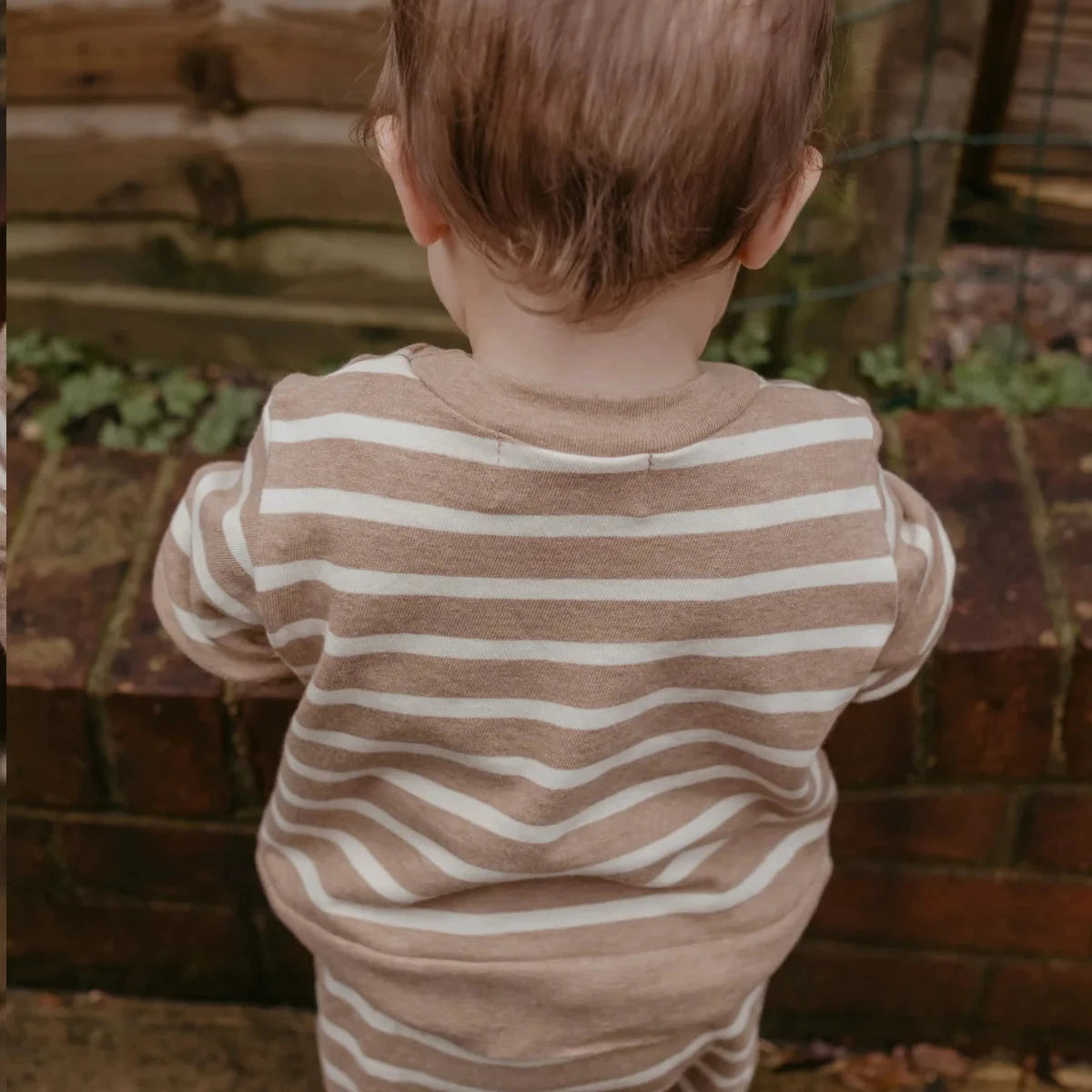 A baby viewed from behind wearing a beige and white horizontally striped long-sleeve romper, standing outdoors in front of a brick wall and wooden fence.