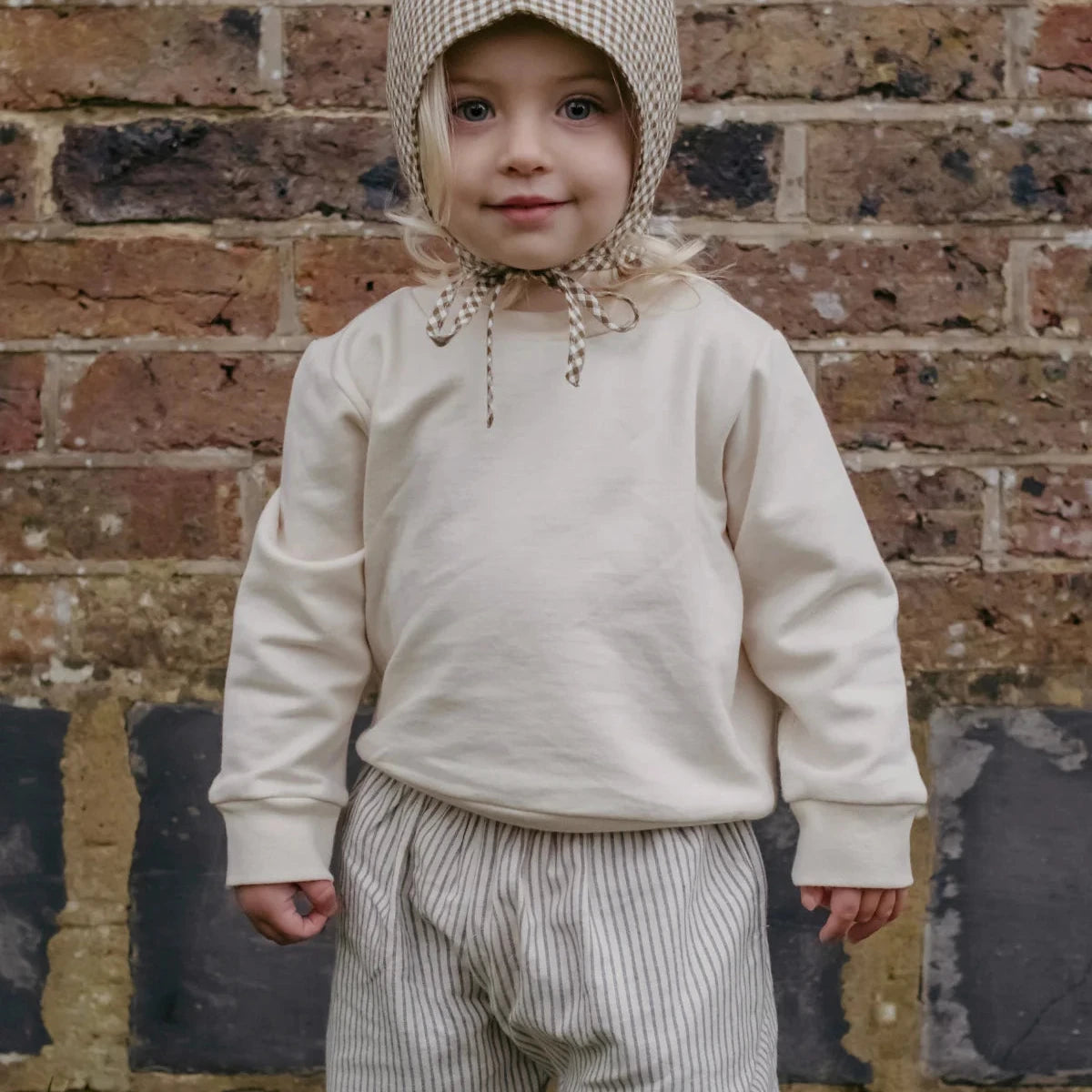 A toddler stands against a rustic brick wall wearing a cream-colored long-sleeve top, matching striped trousers, and a beige-and-white checkered bonnet with chin ties.